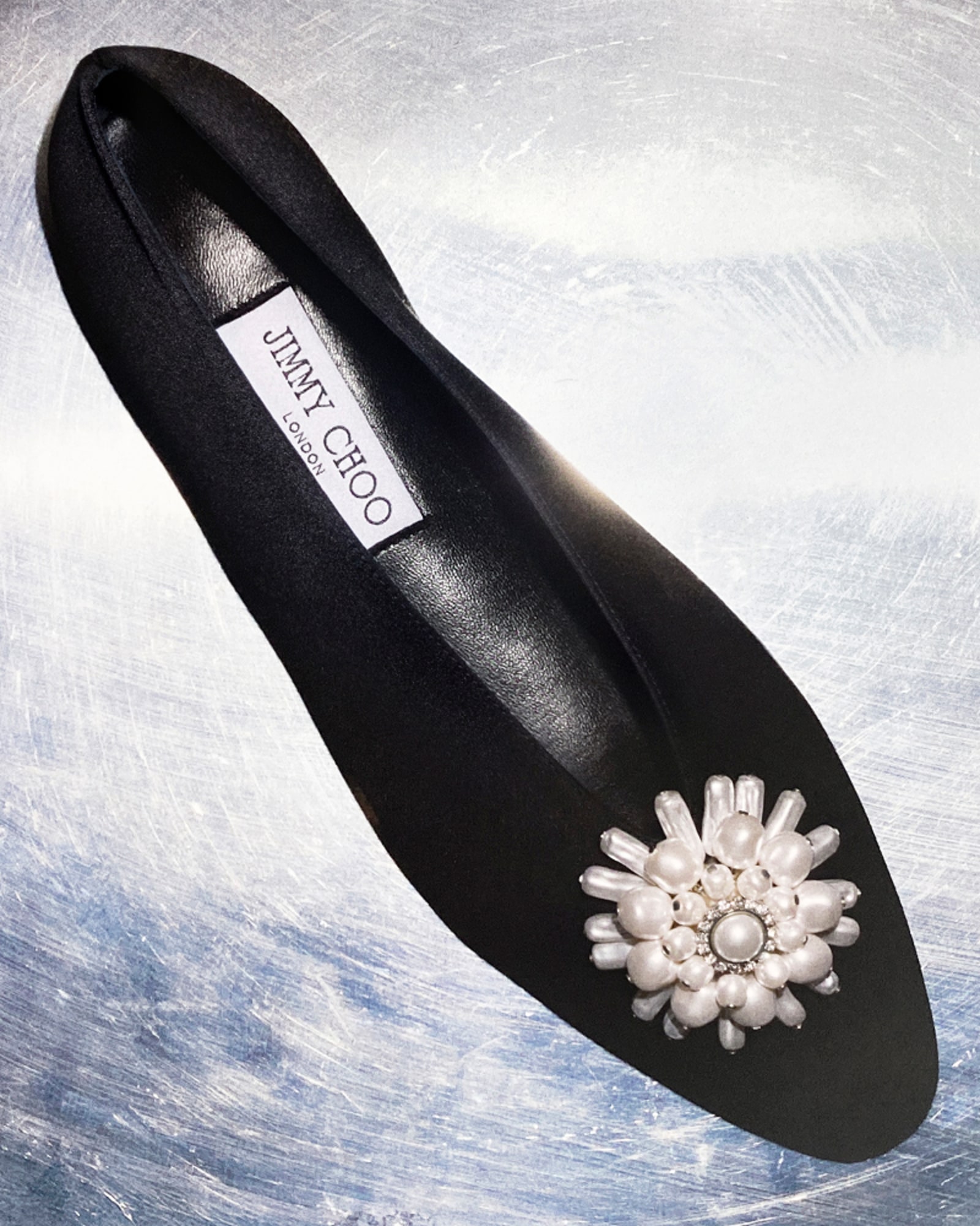 Black satin ballet flat with a pearl and crystal floral embellishment, displayed on a metallic surface.