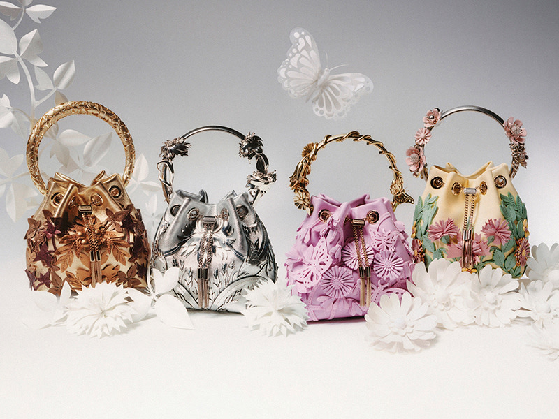 Four Bon Bon bucket bags with floral embellishments and statement bracelet handles in gold, silver, pink and yellow, displayed among white paper flowers.