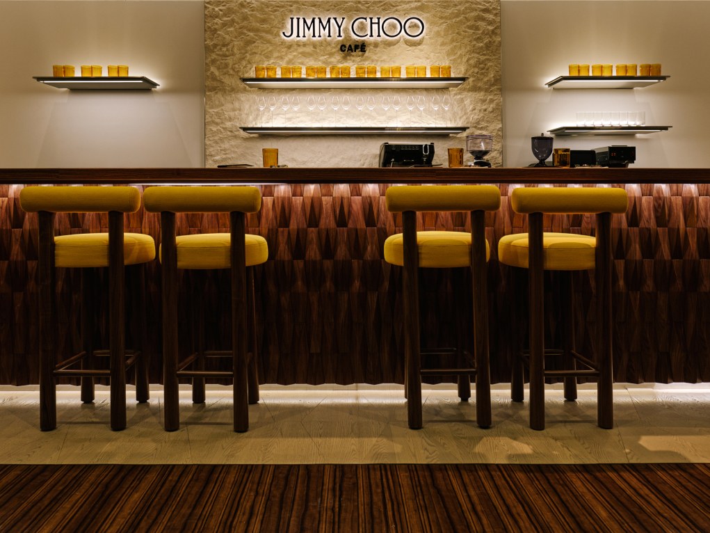 Man seated in a workshop wearing a hat and layered tailoring, with a leather bag placed on a worktable beside him.