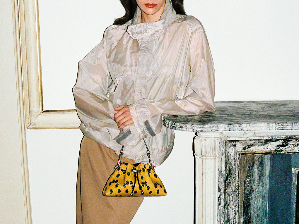 Model wearing a silk jacket and tan skirt, holding a yellow and black spotted mini drawstring bag.