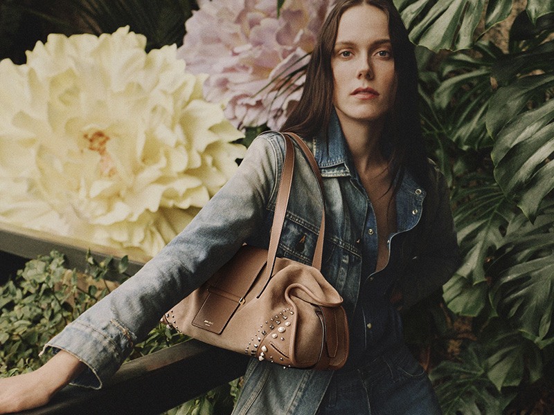 Woman wearing a denim jacket and carrying a tan leather shoulder bag, standing in front of large floral arrangements and green foliage.