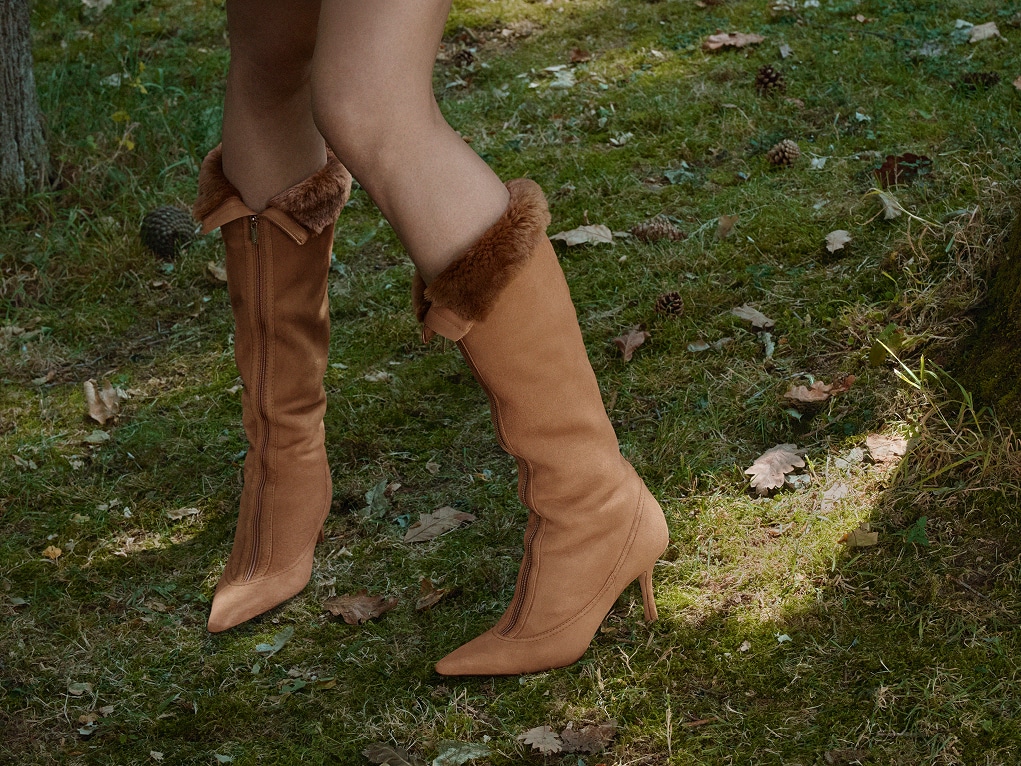  Close-up of tan suede knee-high boots with shearling trim and pointed toes, worn outdoors on grass.