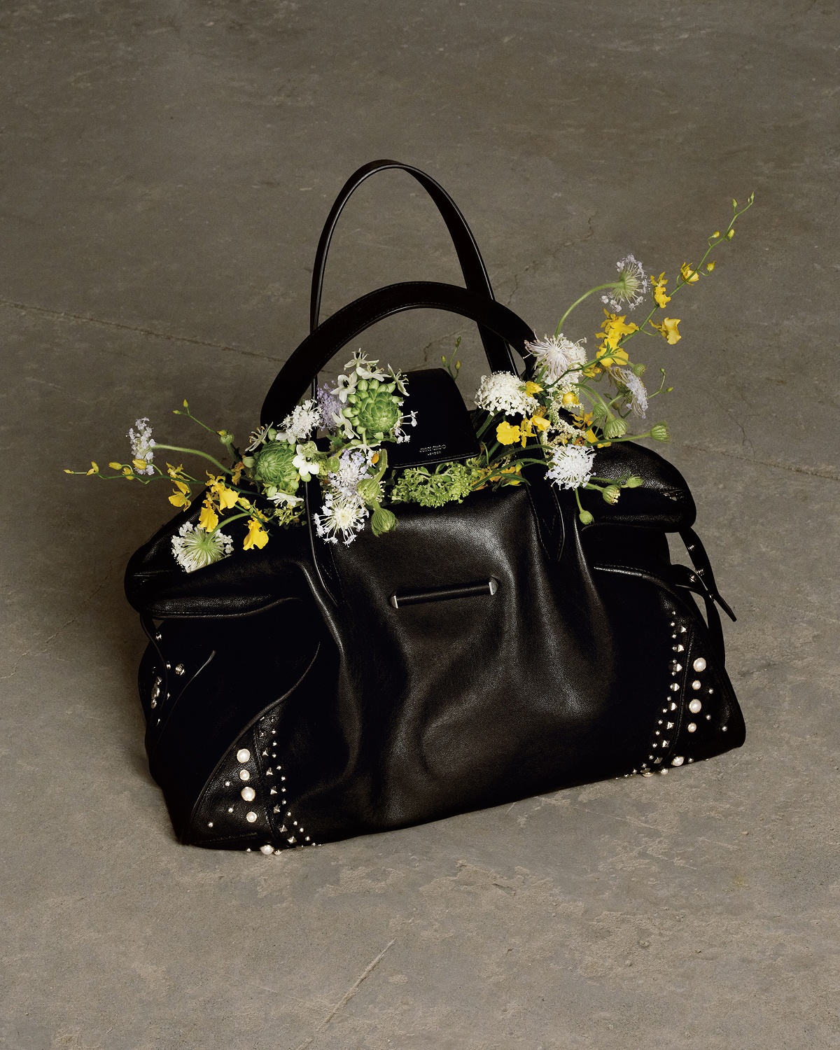 Black leather tote bag with top handles displayed on a concrete plinth outdoors, with bonsai trees in the background.