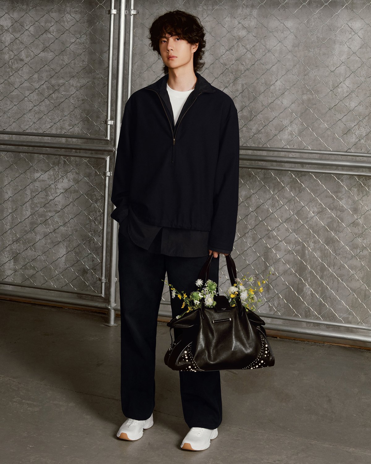 Man seated in a workshop wearing a hat and layered tailoring, with a leather bag placed on a worktable beside him.
