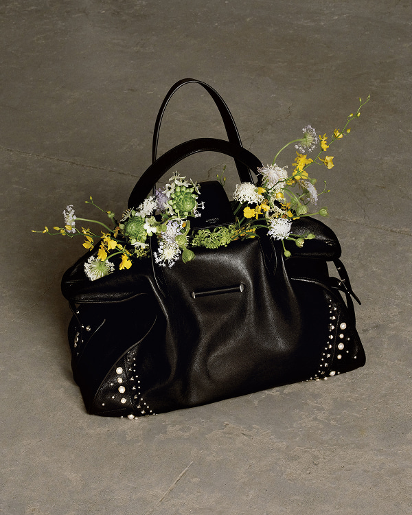 Black leather tote bag with top handles displayed on a concrete plinth outdoors, with bonsai trees in the background.