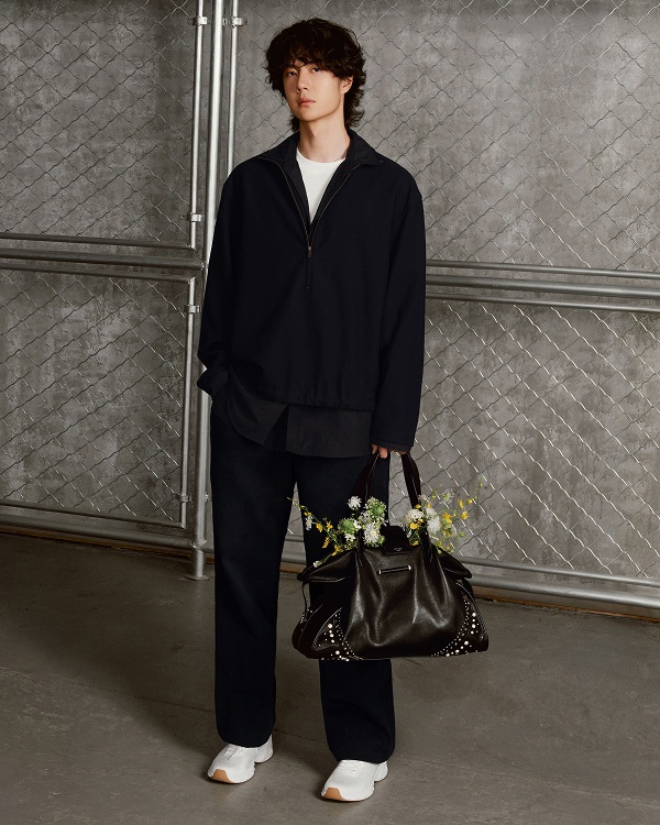 Man seated in a workshop wearing a hat and layered tailoring, with a leather bag placed on a worktable beside him.