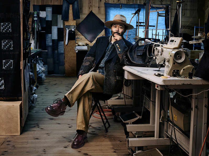 Man seated in a workshop wearing a hat and layered tailoring, with a leather bag placed on a worktable beside him.