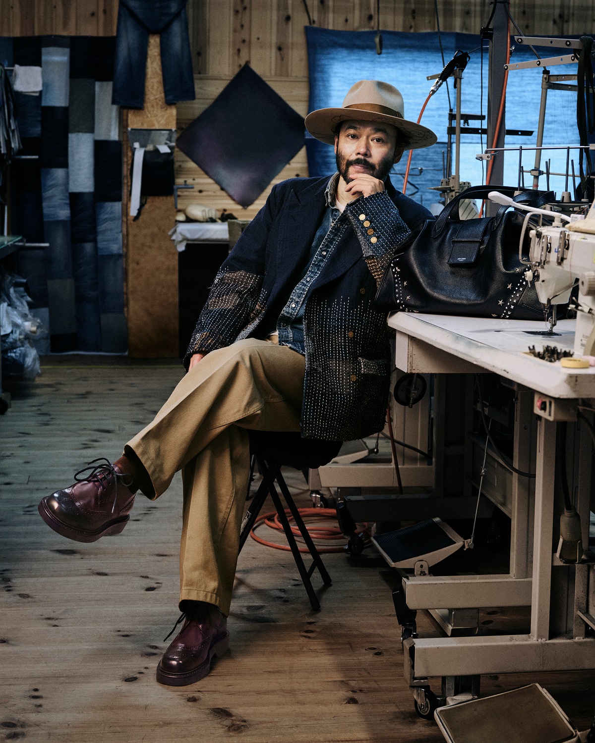 Man seated in a workshop wearing a hat and layered tailoring, with a leather bag placed on a worktable beside him.